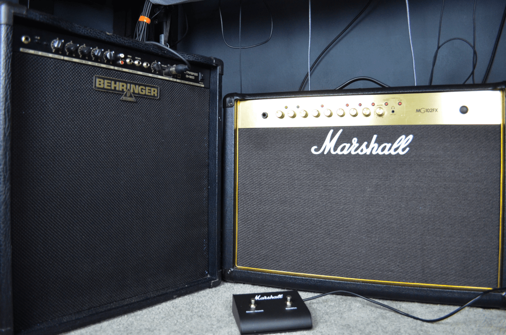 Left half of the image shows a Behringer bass amplifier, right side features a Marshall guitar amplifier. The underside of a desk is visible above