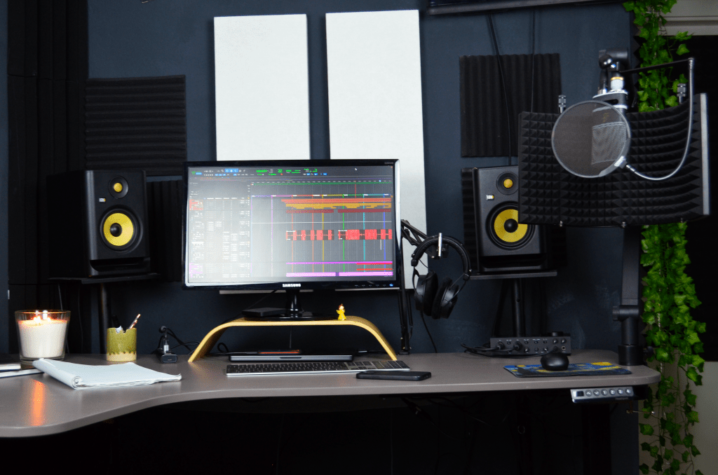 The top half of the image shows a computer screen with white soundproofing panels behind it. The bottom half displays a desk with a laptop. On the right side, a KRK speaker is visible in the middle.