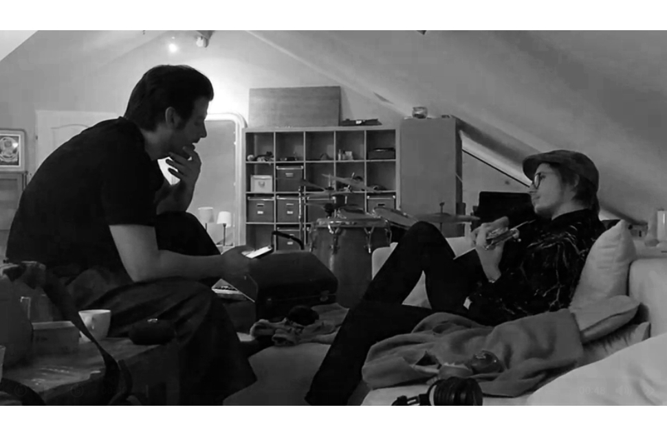 Two musicians relaxing in a studio: one sitting forward on a chair to the right, the other leaning back on a couch with a guitar.